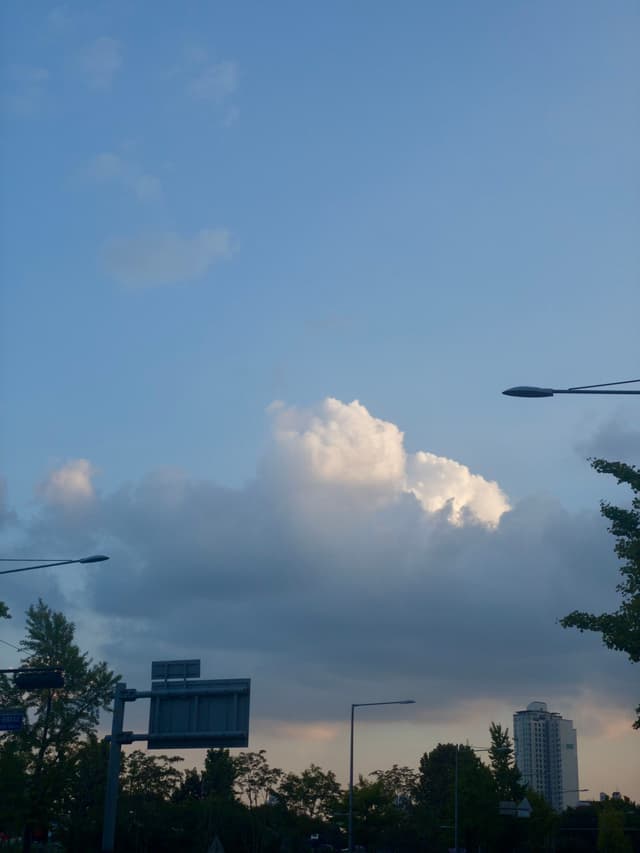 A cityscape with a clear blue sky and fluffy clouds, featuring streetlights, trees, and a tall building in the background