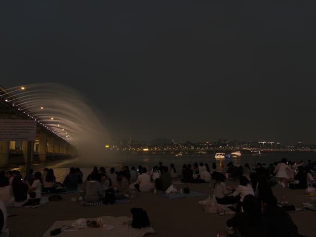 Crowd seated along a riverside promenade at night with a curved canopy structure and city lights reflecting on the water