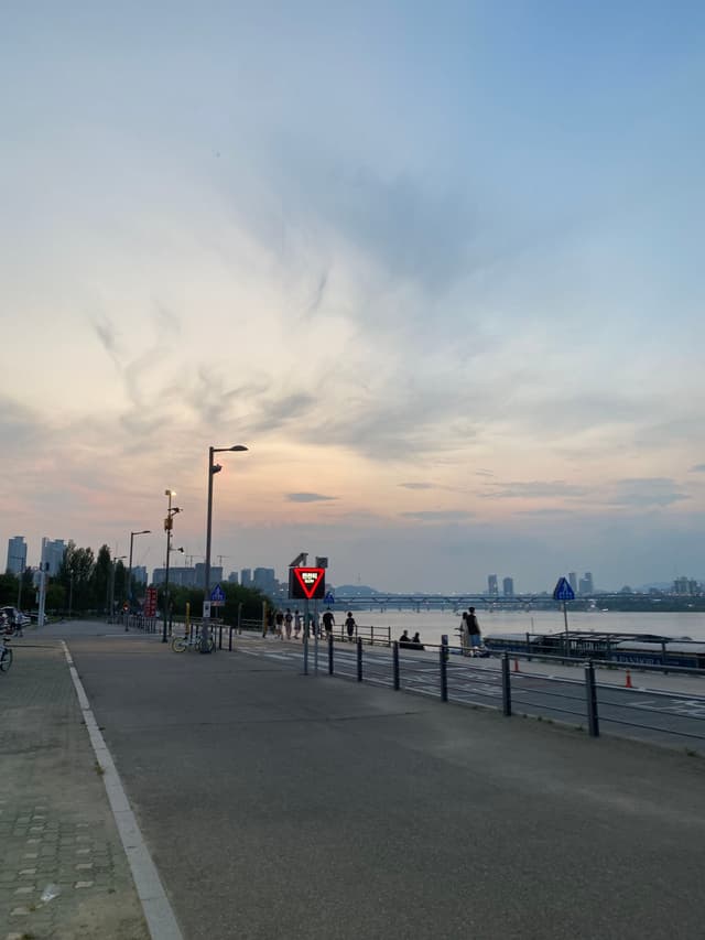 A riverside walkway at dusk with a few people strolling, streetlights, and a distant city skyline under a cloudy sky
