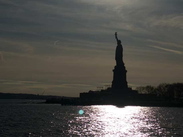 A silhouette of the Statue of Liberty against a darkening sky, with sunlight reflecting on the water
