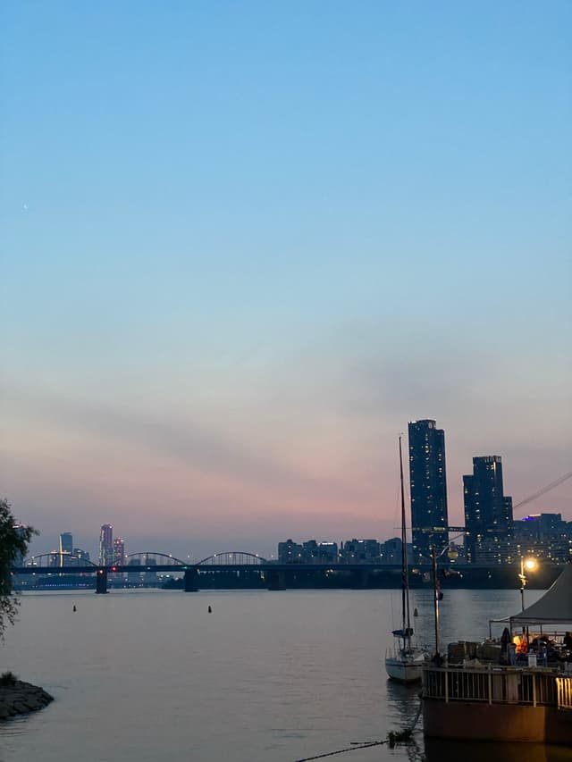 A waterfront cityscape at dusk with tall buildings, a bridge, and boats docked along the shore under a clear sky transitioning from blue to pink