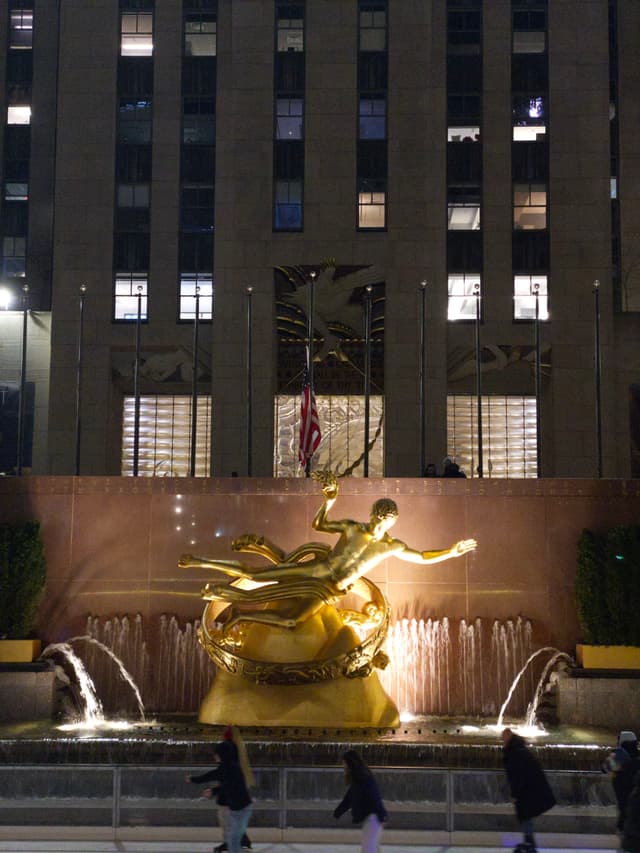 A golden statue of Prometheus is set against a backdrop of a building with illuminated windows, surrounded by a fountain and people skating in the foreground