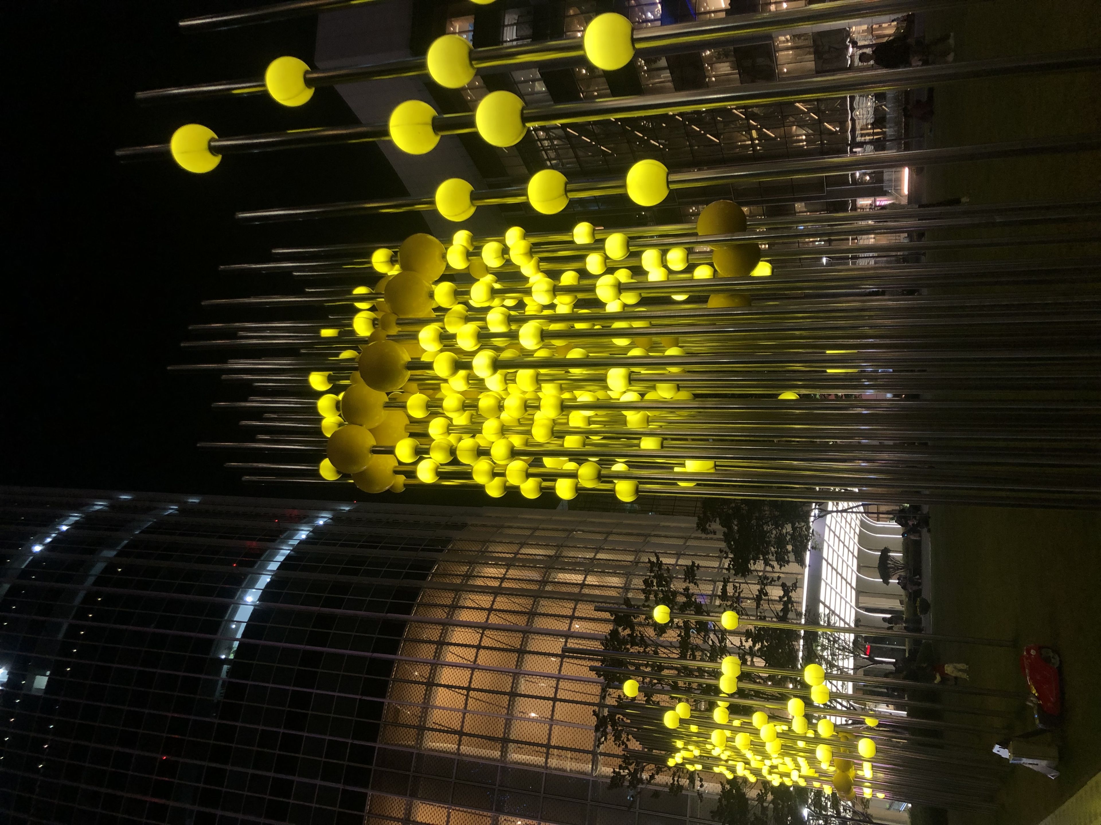 A nighttime scene featuring tall, illuminated yellow spheres on poles, set against a backdrop of modern buildings