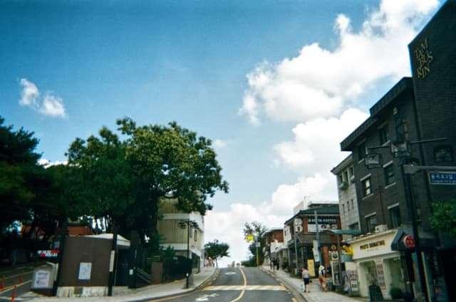 Curving two-lane street through a small town with storefront buildings, trees, and a bright cloudy sky