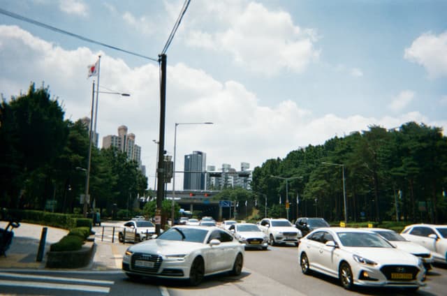 Sedans driving on a multi-lane roadway with trees and a distant skyline under clouds