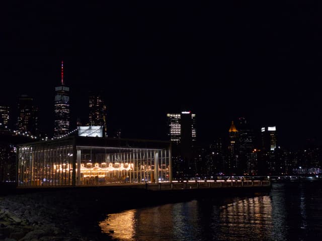A nighttime cityscape featuring illuminated buildings along a waterfront, with reflections on the water and a prominent lit structure in the foreground