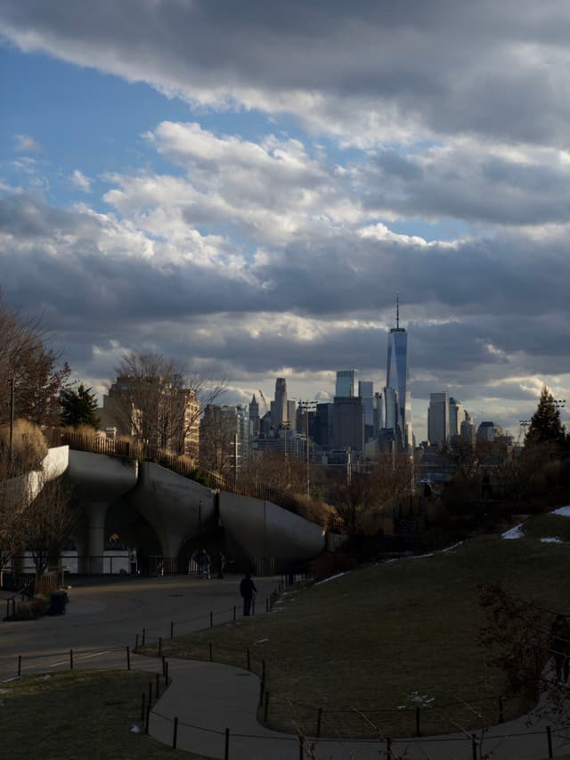A city skyline with tall buildings under a cloudy sky, featuring a park in the foreground with a winding path and a modern bridge structure