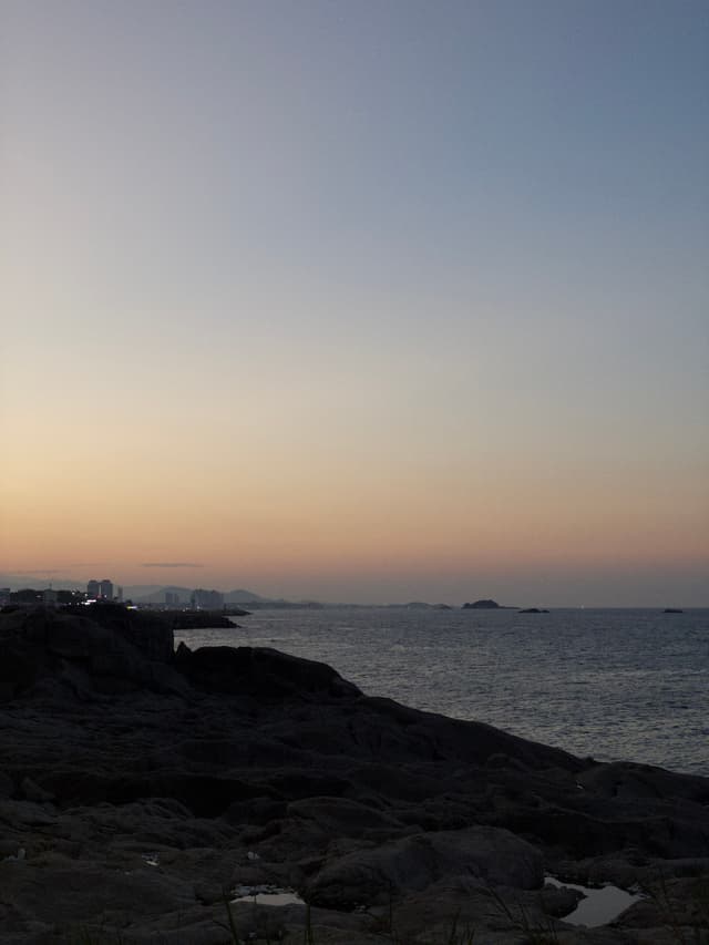 A rocky coastline at sunset, featuring a calm sea under a gradient sky transitioning from orange to blue