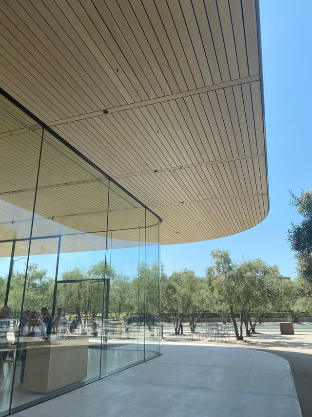 A modern building with large glass walls and a wooden overhang, surrounded by trees under a clear blue sky
