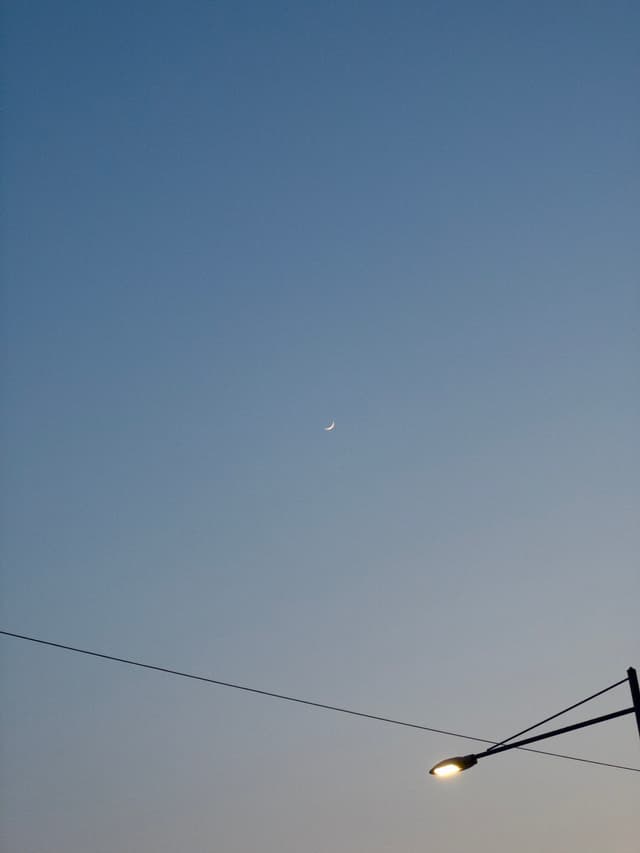 A crescent moon in a clear evening sky with a streetlight and power lines in the foreground