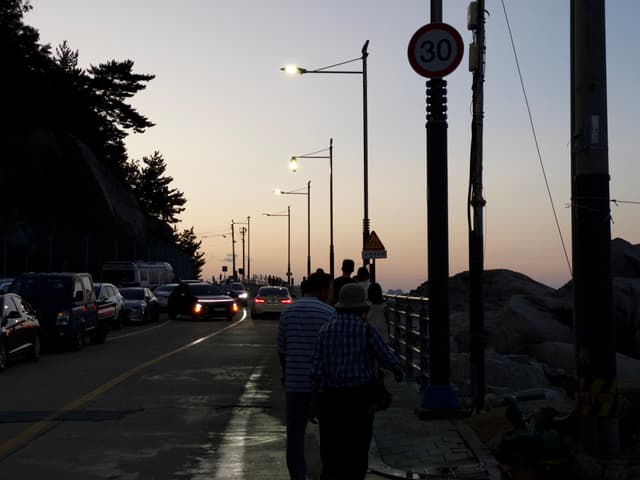 A seaside road at dusk with people walking, cars parked alongside, and streetlights illuminating the path. A 50 km/h speed limit sign is visible, and distant hills are silhouetted against the evening sky
