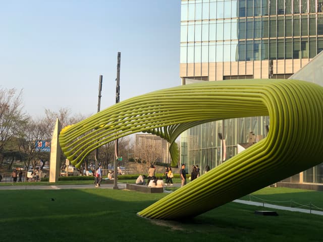 A large, modern yellow sculpture with a curved, ribbed design is situated on a grassy area near a glass building. People are walking nearby, and trees are visible in the background