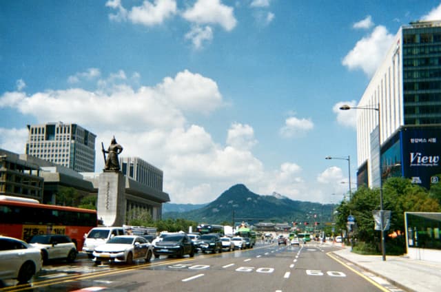 Multi-lane city street with crosswalk, cars, buses, tall buildings, and distant mountains under a partly cloudy sky