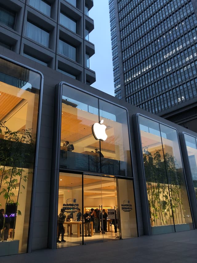 An Apple store with large glass windows, displaying the Apple logo, surrounded by tall buildings