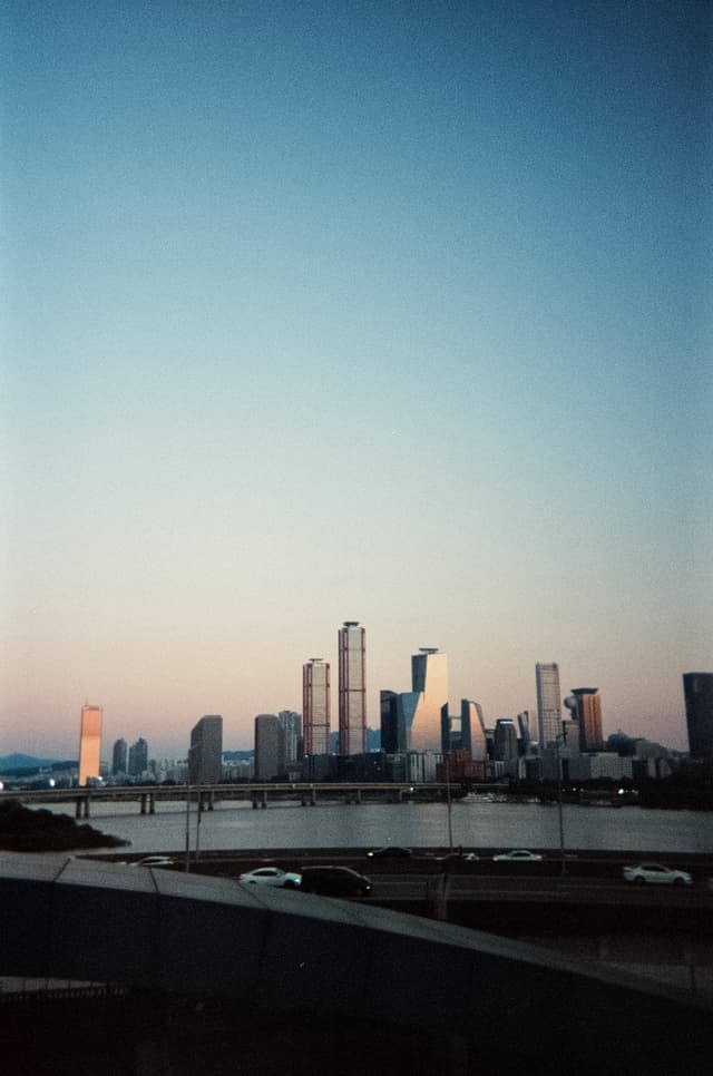 Distant urban skyline with high-rise towers across a river at dusk, viewed from a bridge railing under a clear gradient sky
