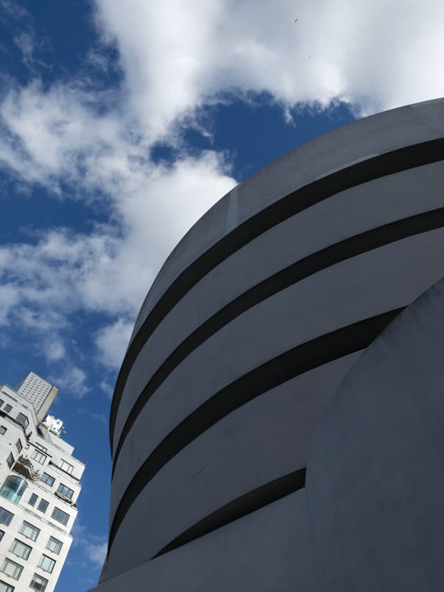 A modern, curved building with horizontal lines against a blue sky with clouds, alongside a more traditional building
