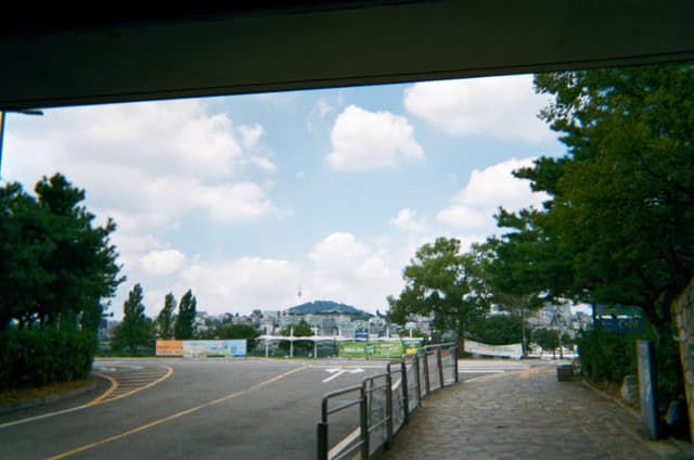 Paved walkway beside a road with railings and trees leading toward a distant bridge along a river under a partly cloudy sky