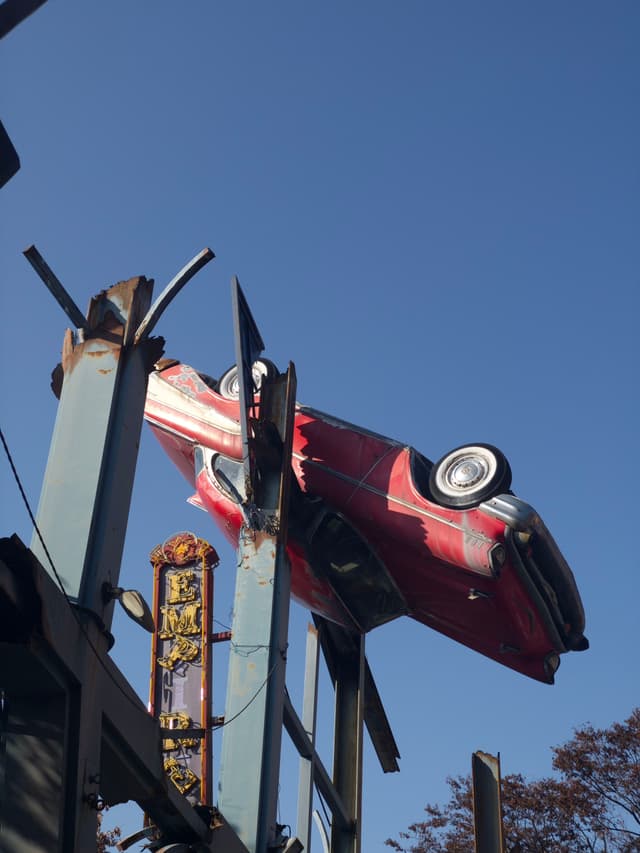 A red vintage car is mounted upside down on metal beams against a clear blue sky
