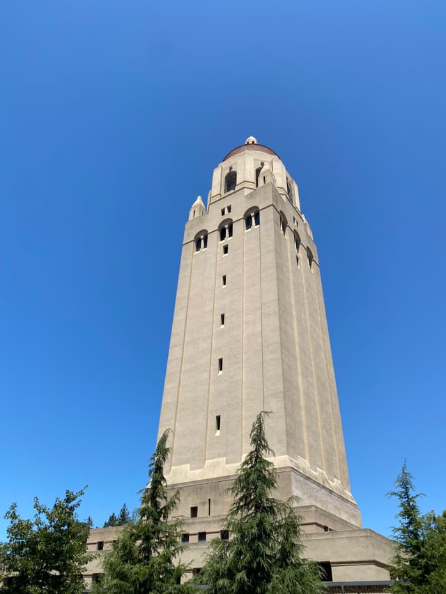 A tall, beige tower with a red-tiled roof set against a clear blue sky, surrounded by trees at the base