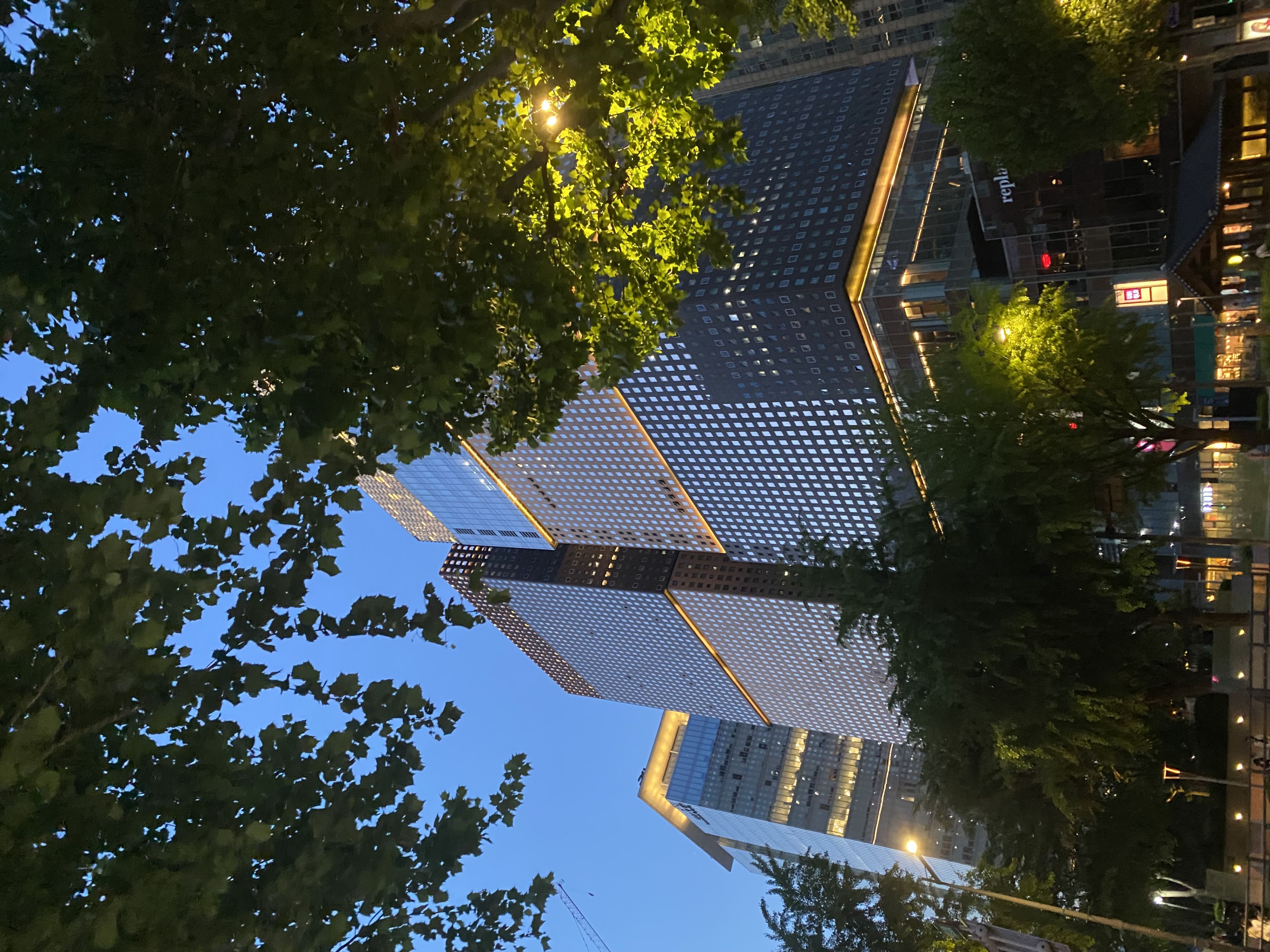 A modern cityscape with tall, illuminated buildings surrounded by trees, set against a twilight sky
