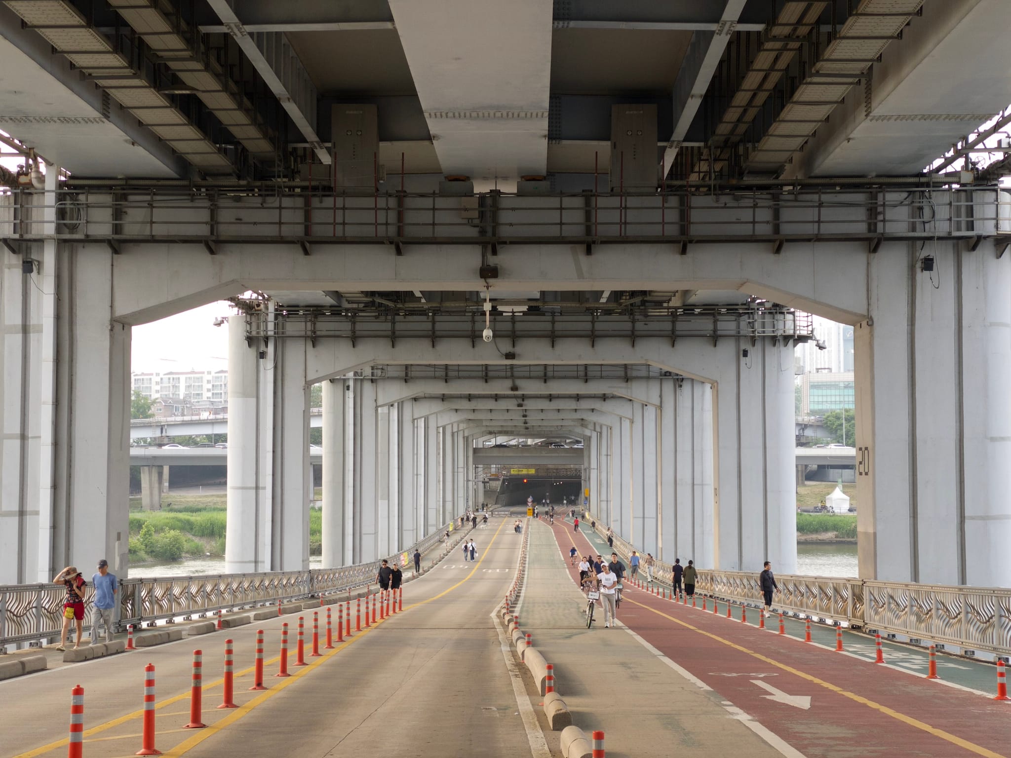 A view under a large bridge with multiple support columns, featuring a road and a separate pedestrian path with people walking