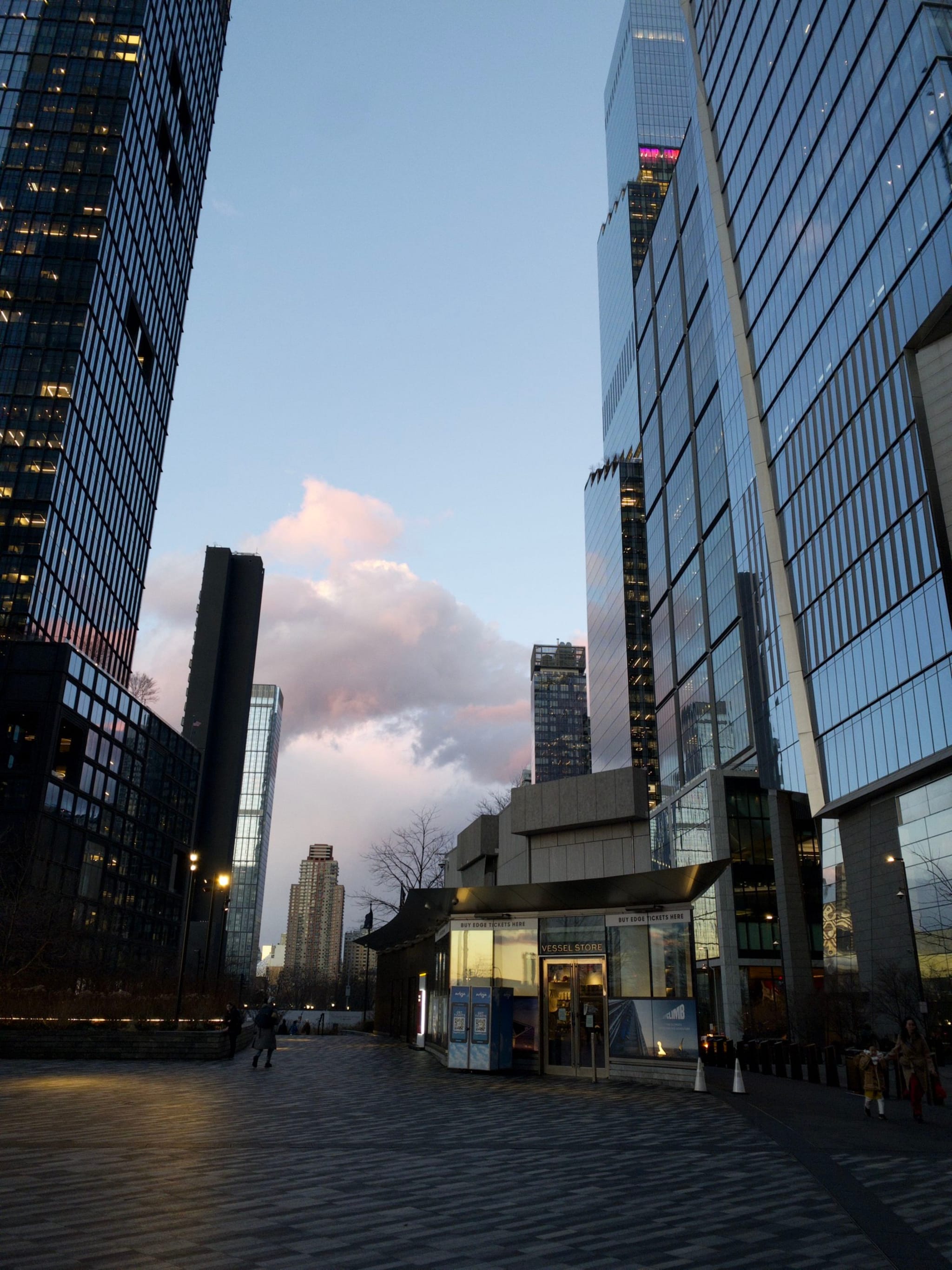 A cityscape at dusk with tall, reflective skyscrapers lining a wide street, capturing a partly cloudy sky in the background