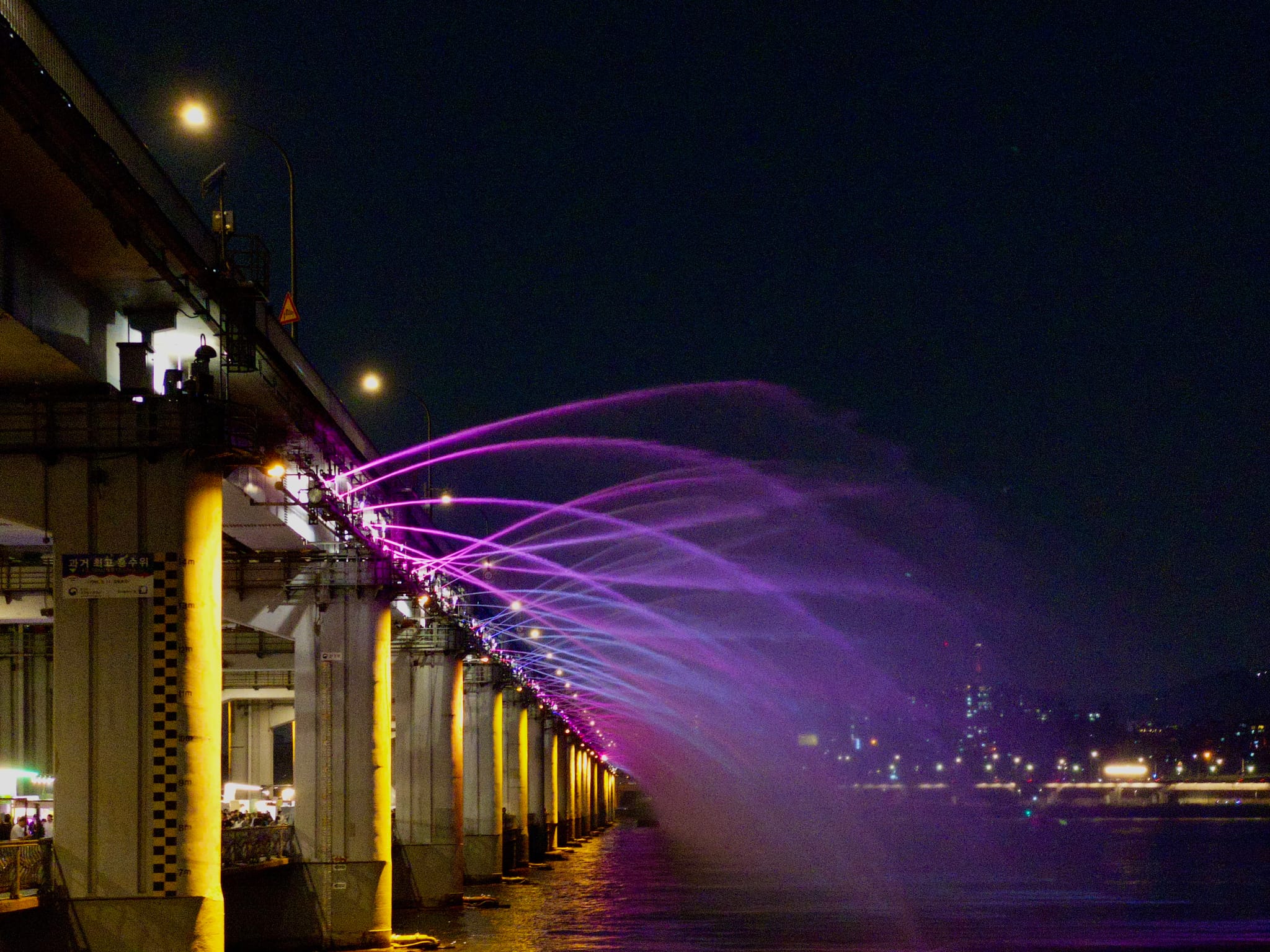 A bridge at night with colorful illuminated water jets arching over the side, creating a vibrant display against the dark sky