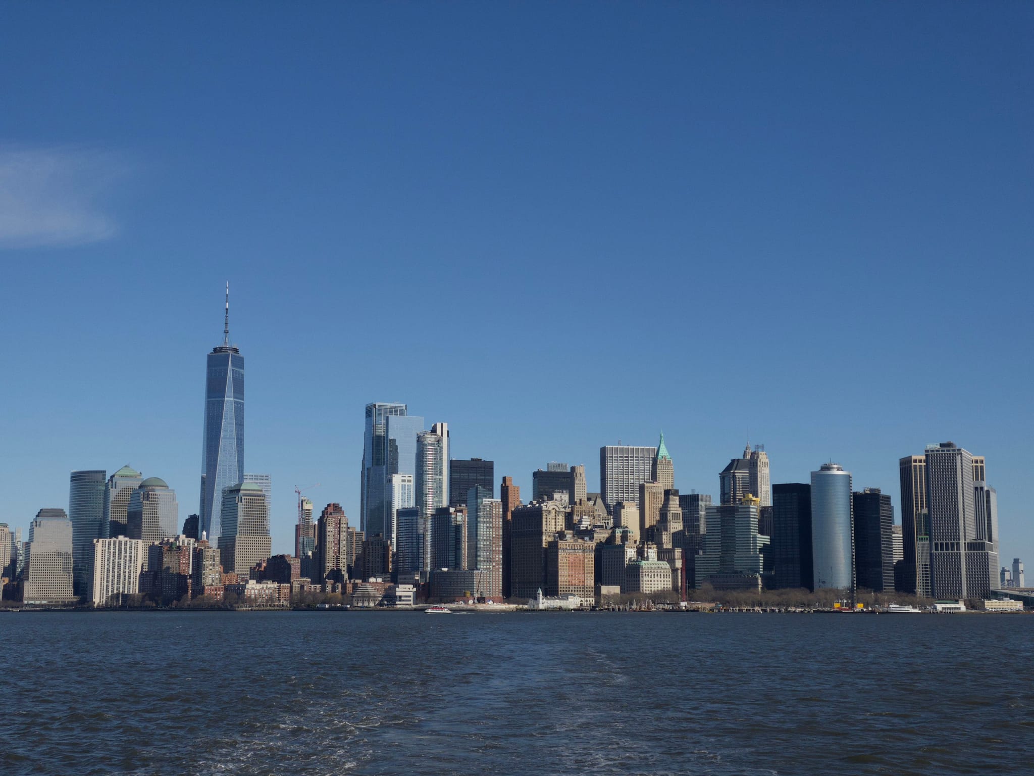 A city skyline with numerous skyscrapers under a clear blue sky, viewed from across a body of water