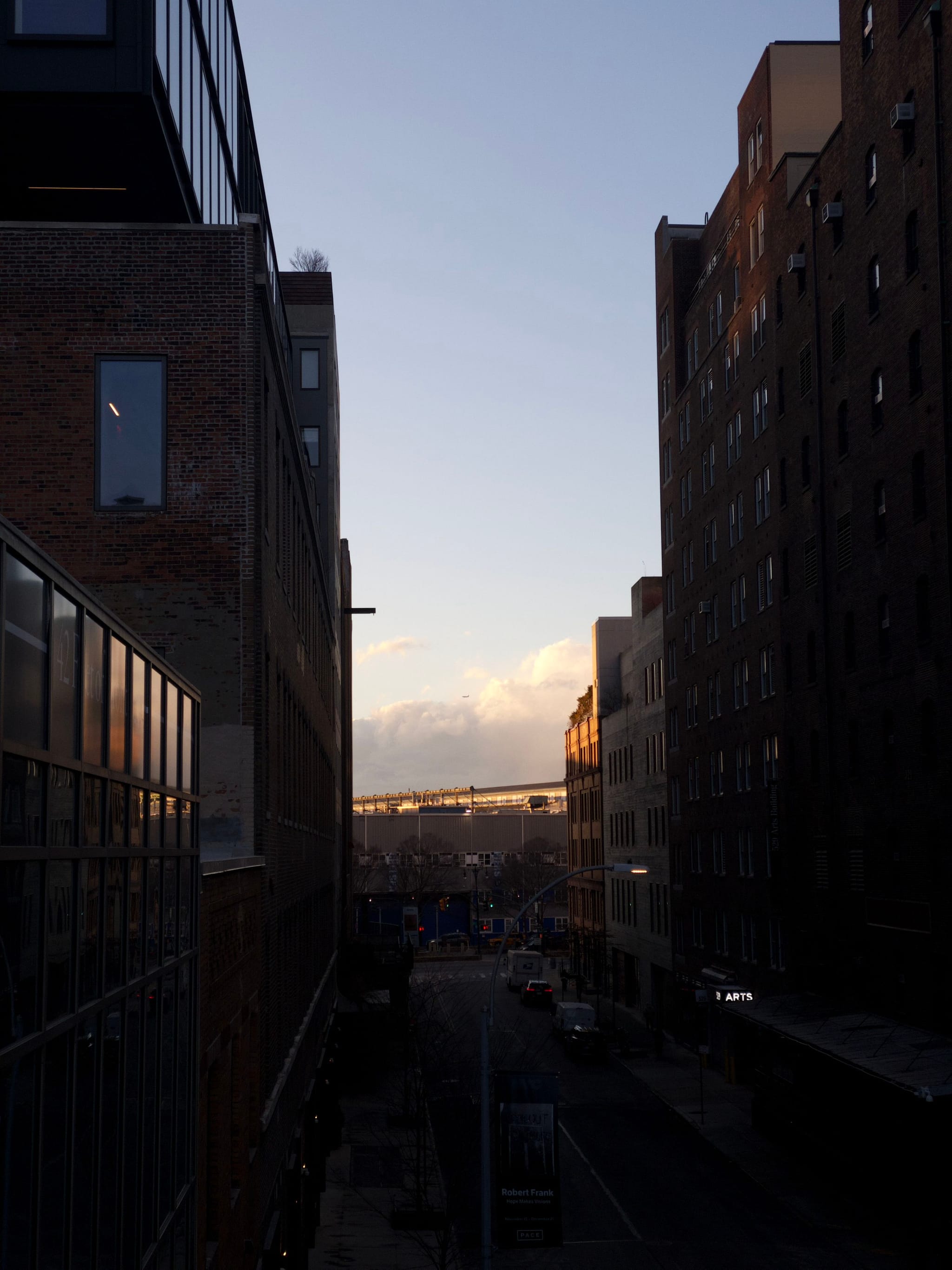 A city street is flanked by tall buildings on both sides, with a view of the sky and clouds in the background, illuminated by the setting sun