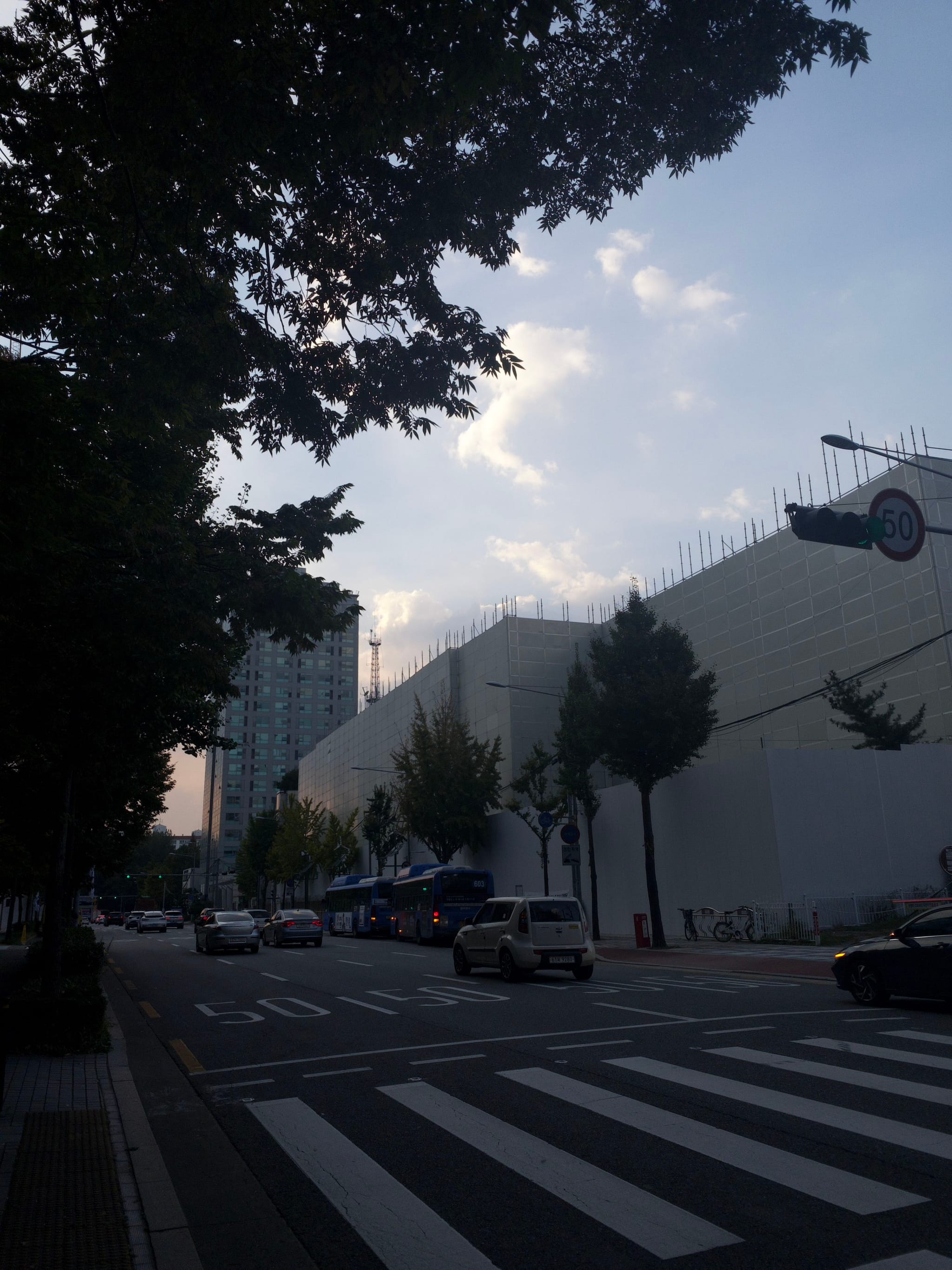 A city street with cars, trees lining the sidewalk, and a large building under construction. The sky is partly cloudy