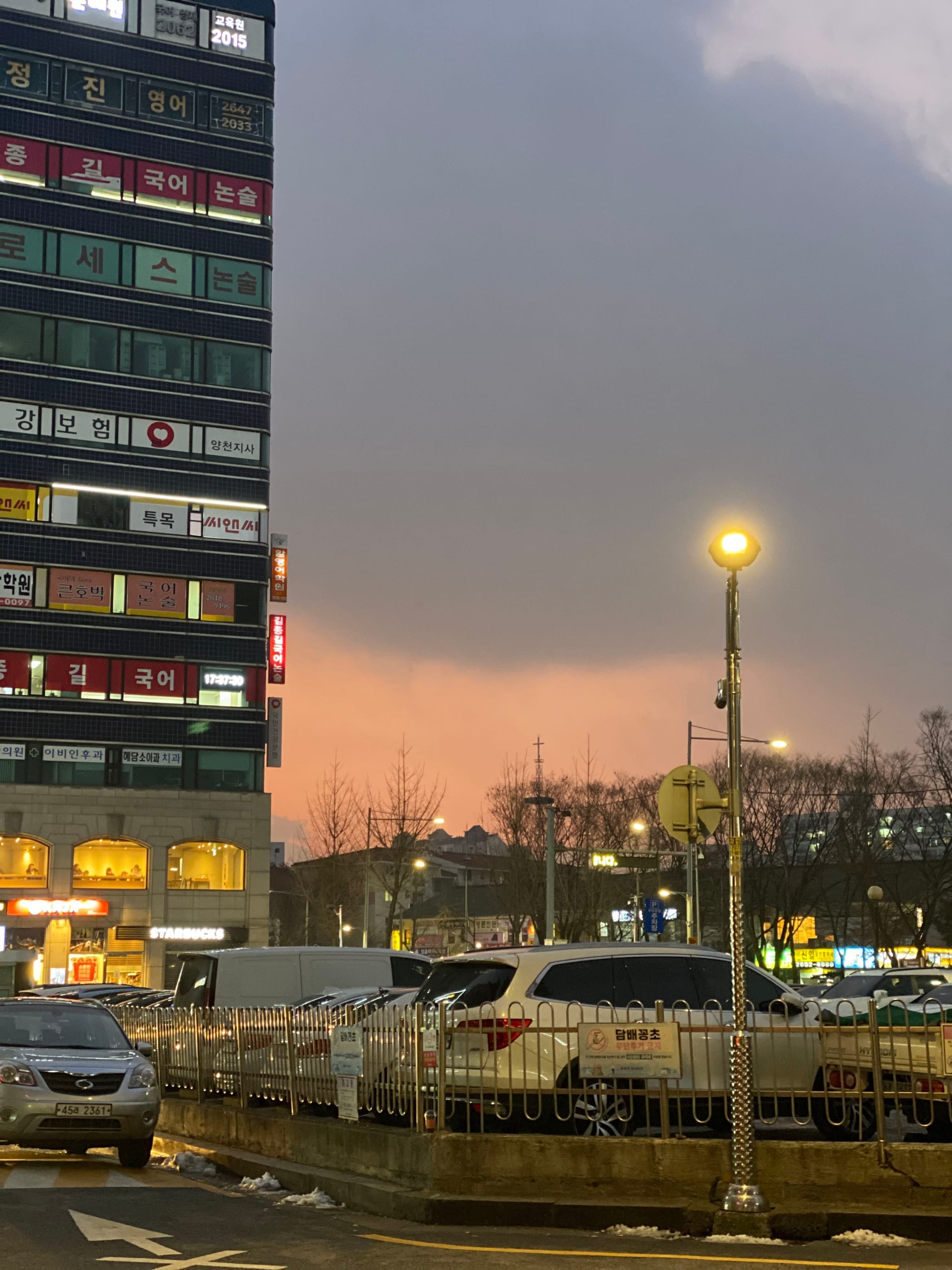 A cityscape at dusk with a tall building on the left, a parking lot with cars in the foreground, and a streetlamp illuminating the scene. The sky is cloudy with a hint of sunset colors near the horizon