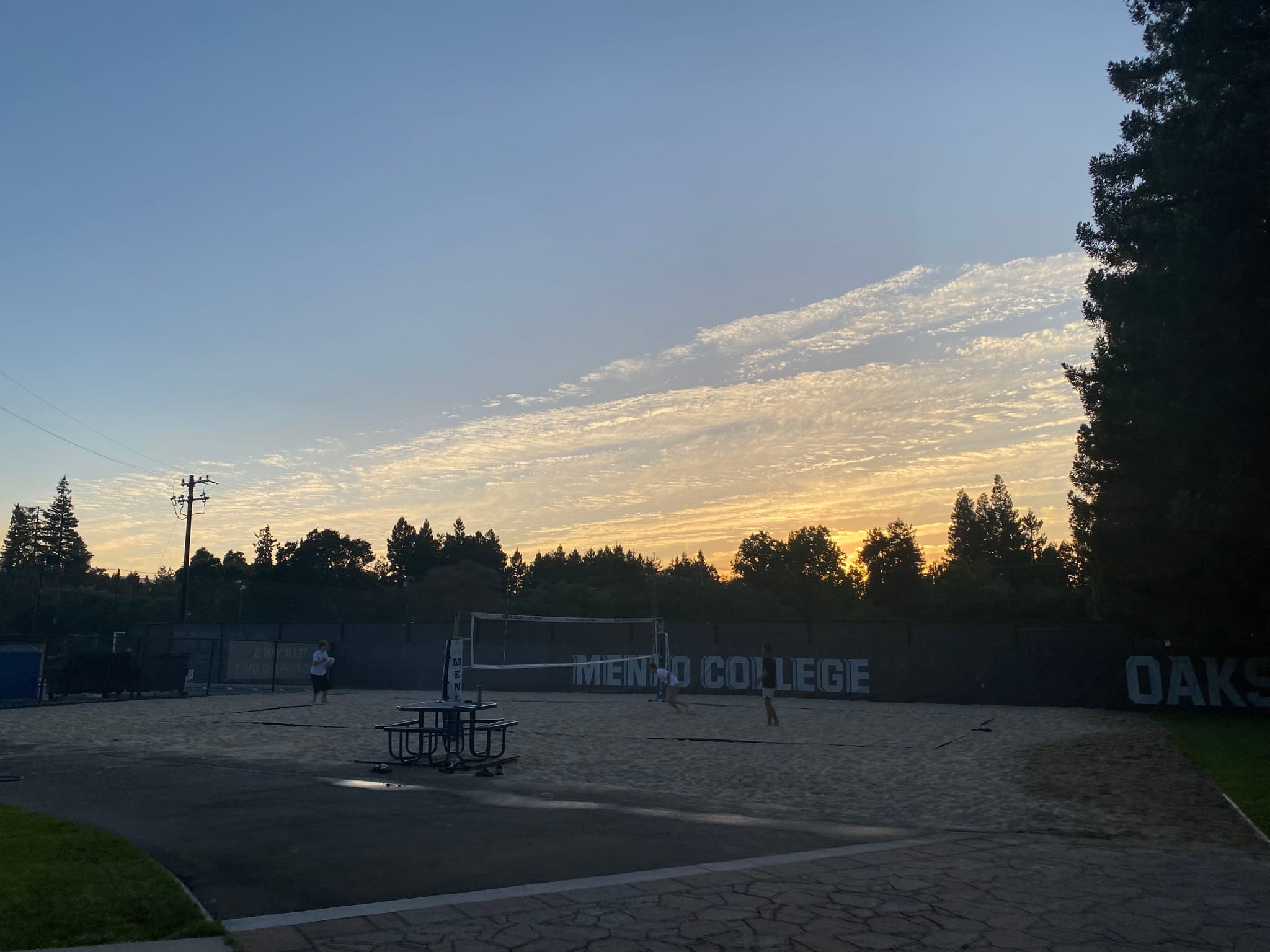 A volleyball court at sunset with a clear sky and scattered clouds, surrounded by trees and a fence with Oaks and Mento College visible