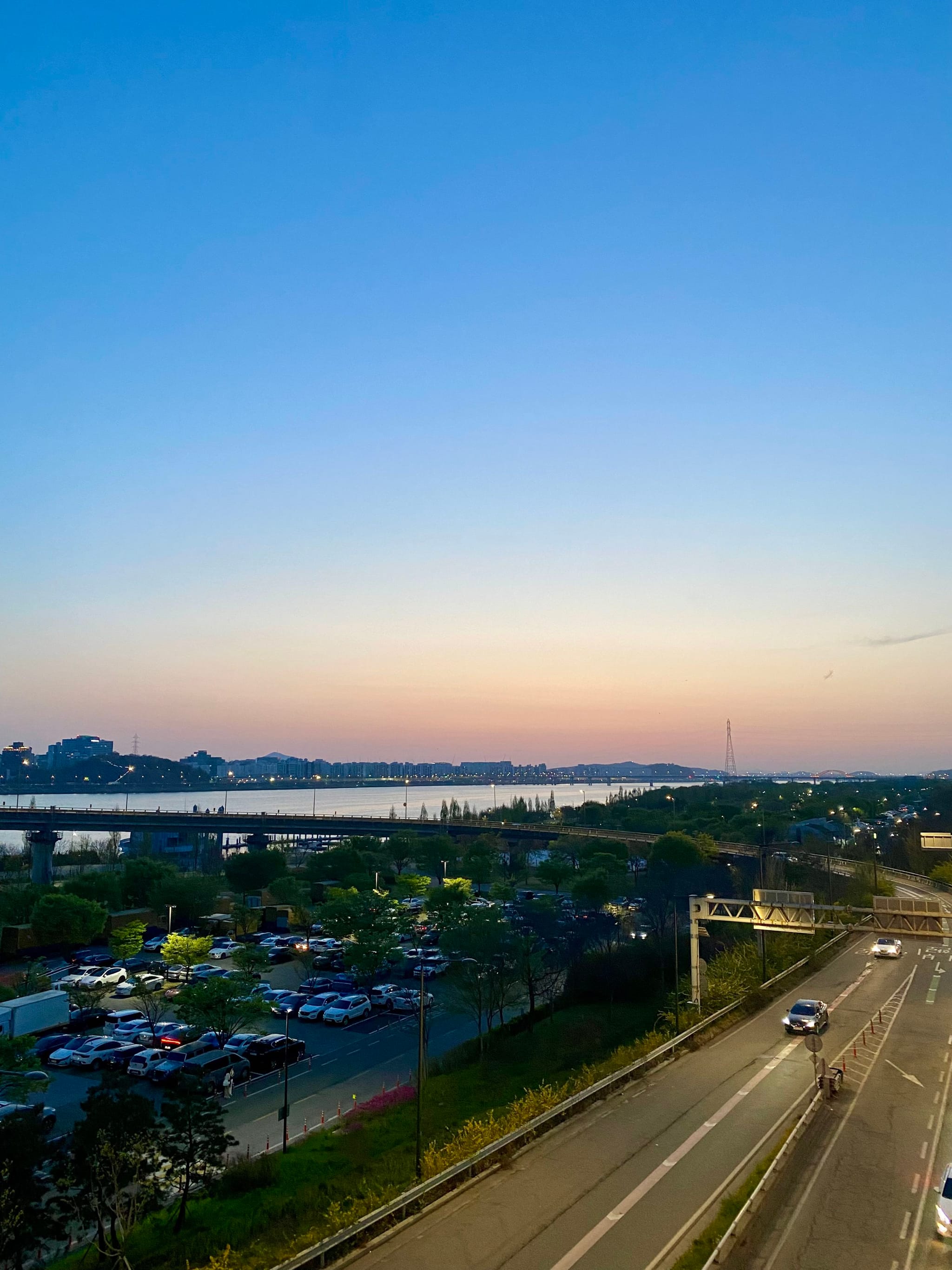 A serene sunset over a river with a clear sky, a parking lot, and a road with light traffic