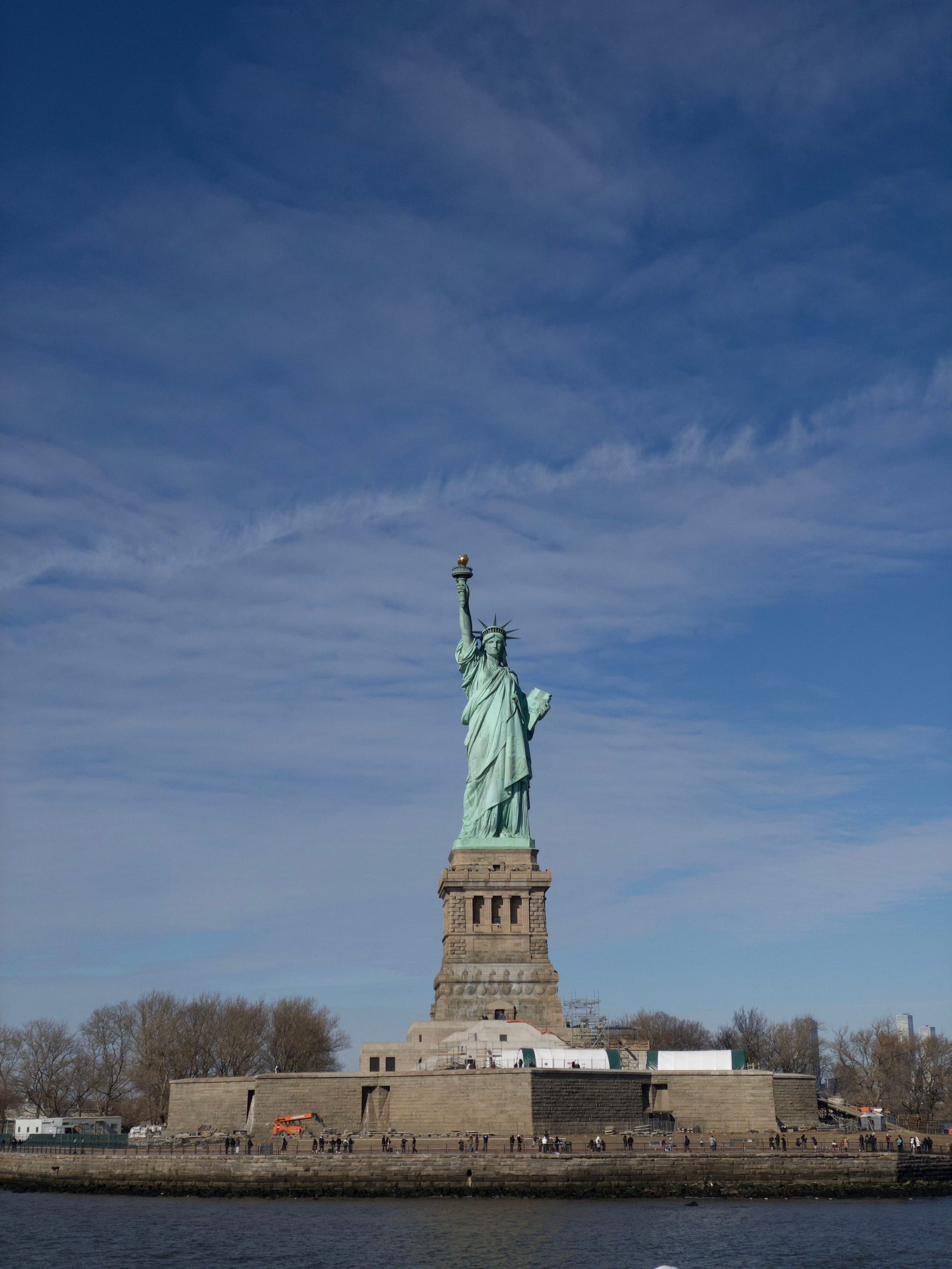 The Statue of Liberty stands on Liberty Island against a backdrop of a partly cloudy sky, with water in the foreground