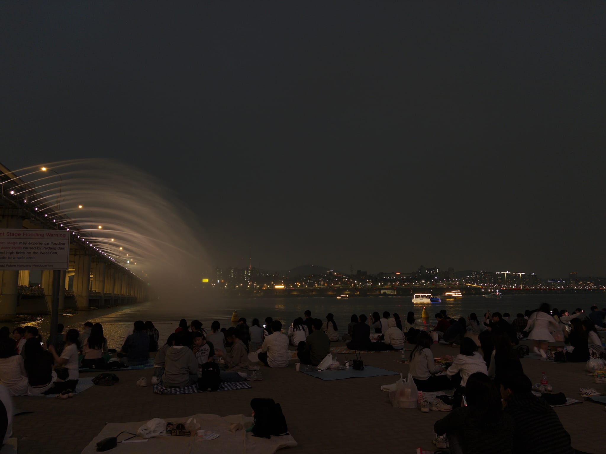 Crowd seated along a riverside promenade at night with a curved canopy structure and city lights reflecting on the water