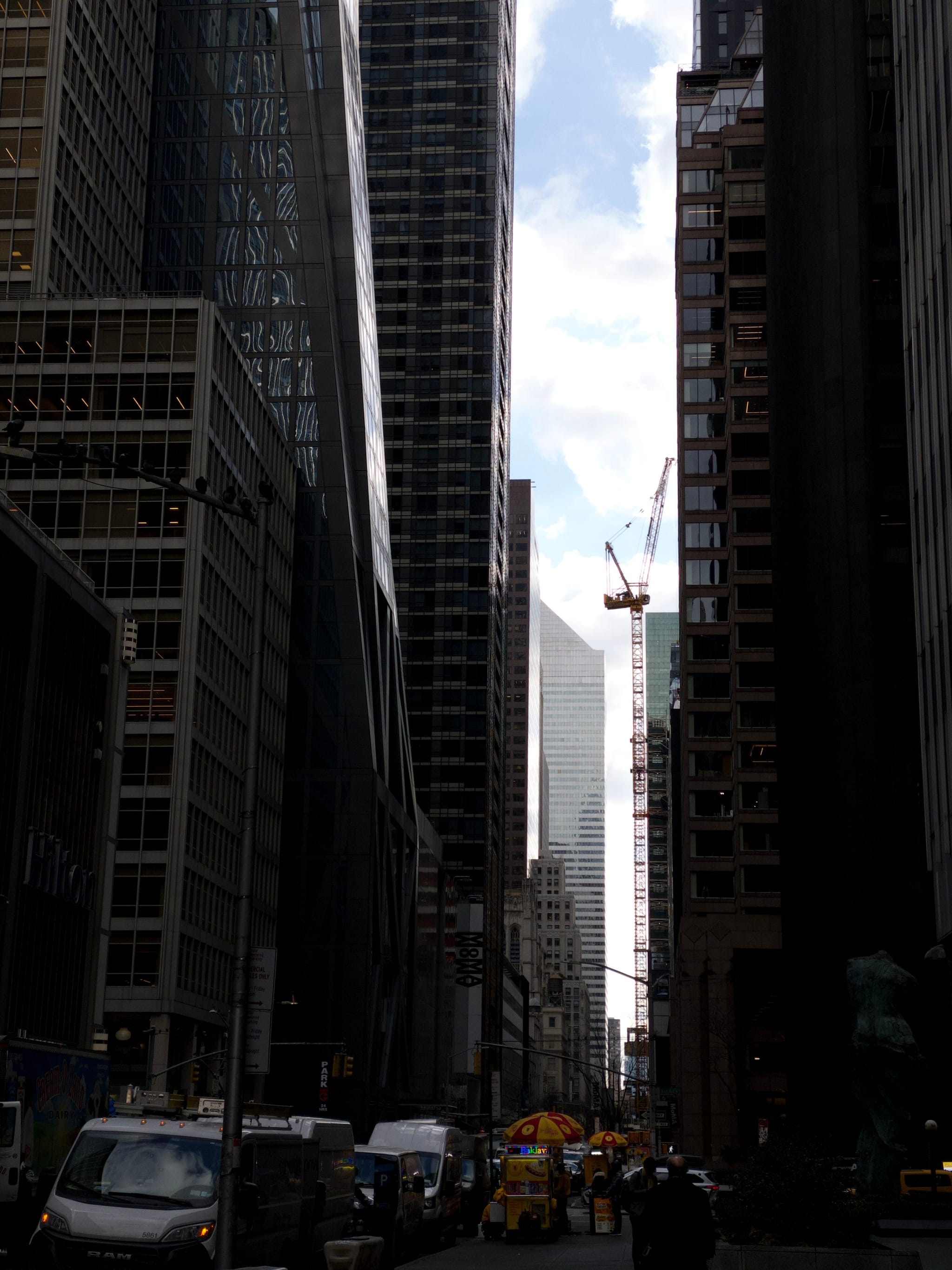 A city street lined with tall skyscrapers, featuring a construction crane in the distance and a food cart on the sidewalk