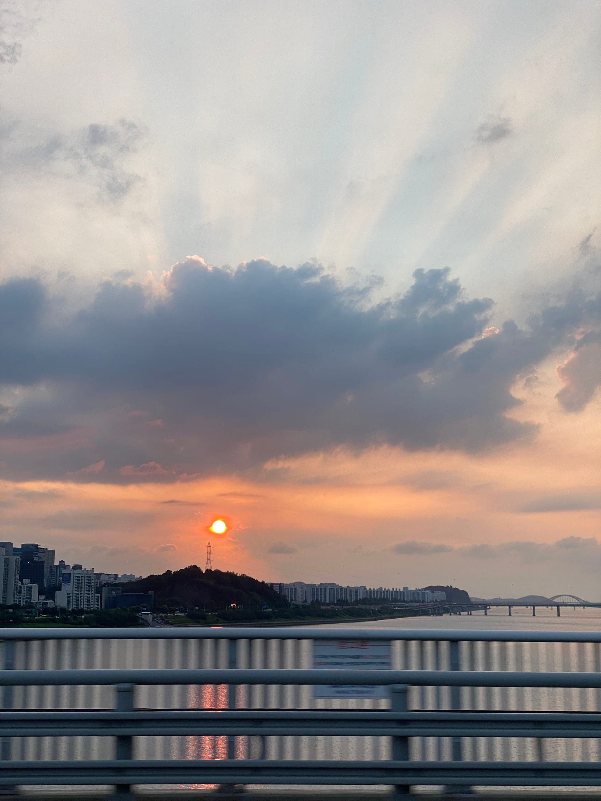 A sunset over a cityscape with a river, bridge, and buildings silhouetted against a sky filled with clouds and sun rays