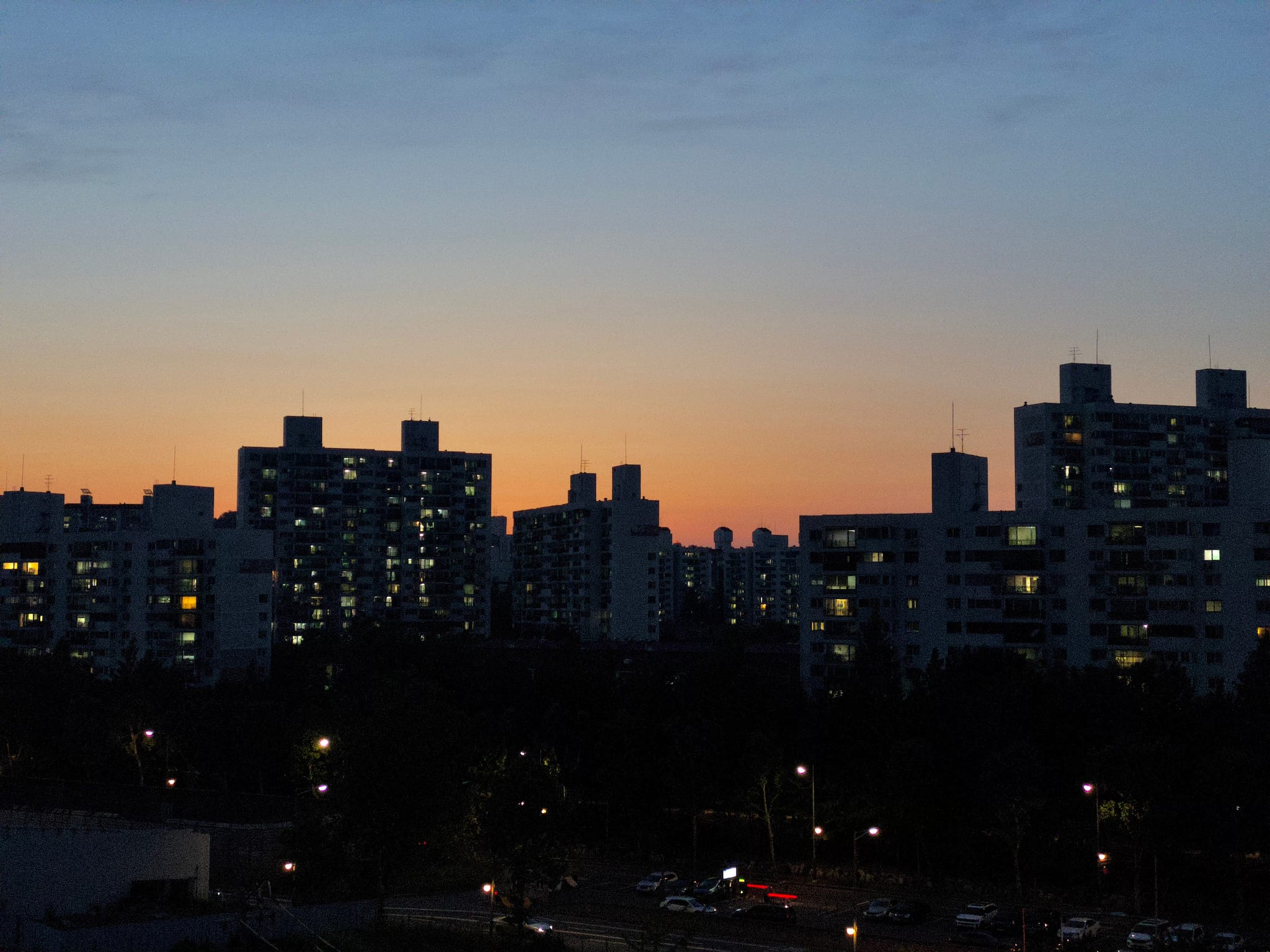 A cityscape at dusk with silhouetted buildings against a colorful sunset sky, featuring illuminated windows and streetlights