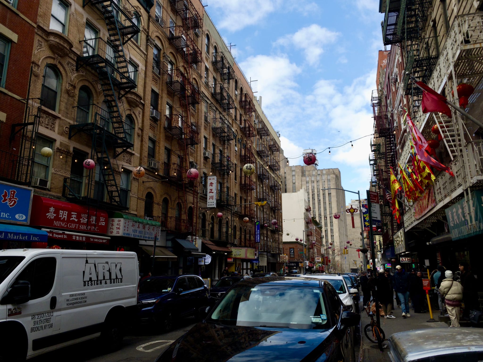 A bustling urban street scene with parked cars, pedestrians, and buildings adorned with fire escapes and colorful signs. Lanterns and flags hang above the street, and the sky is partly cloudy