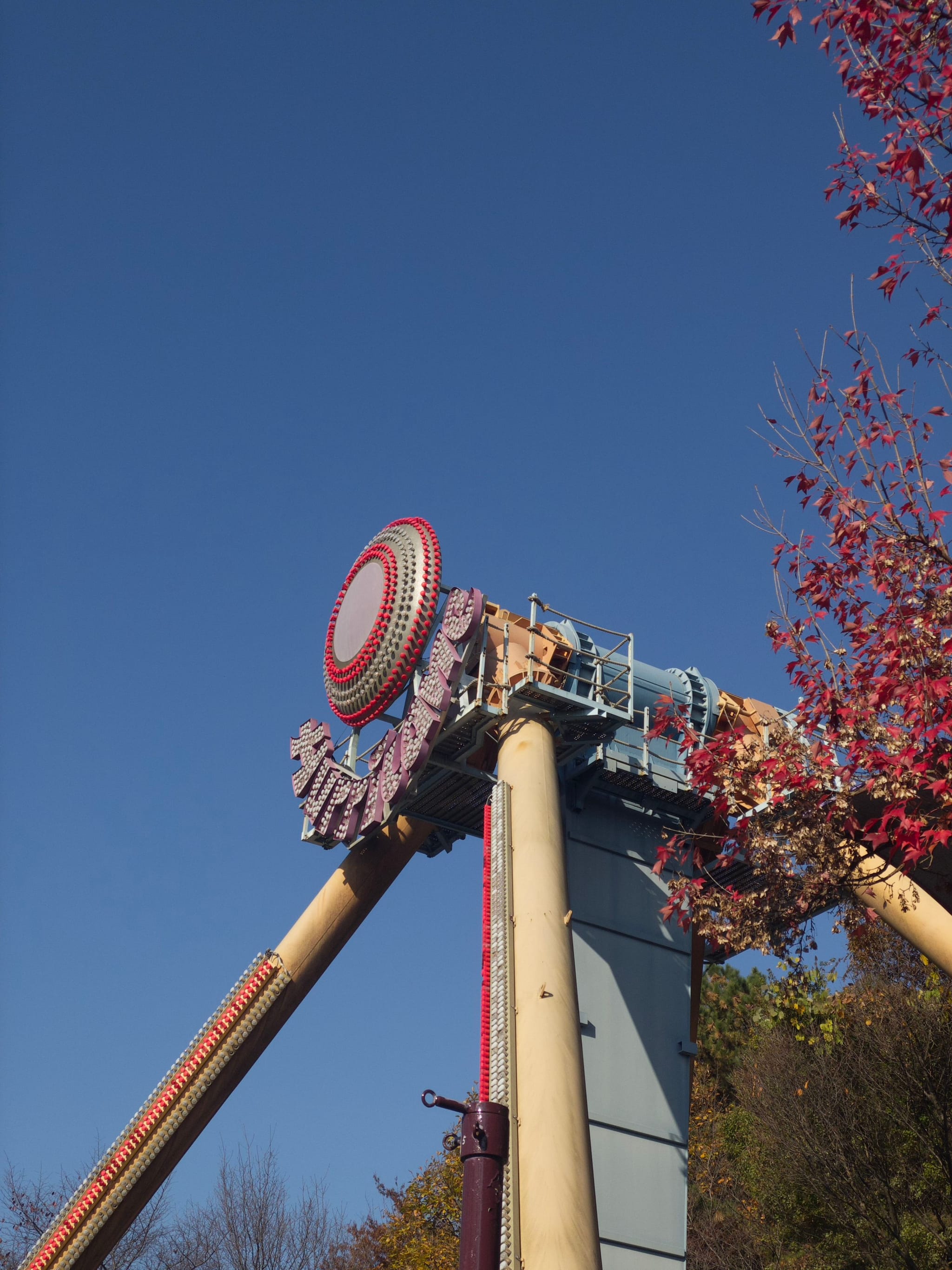 A large amusement park ride with a circular design at the top, set against a clear blue sky and surrounded by trees with autumn foliage