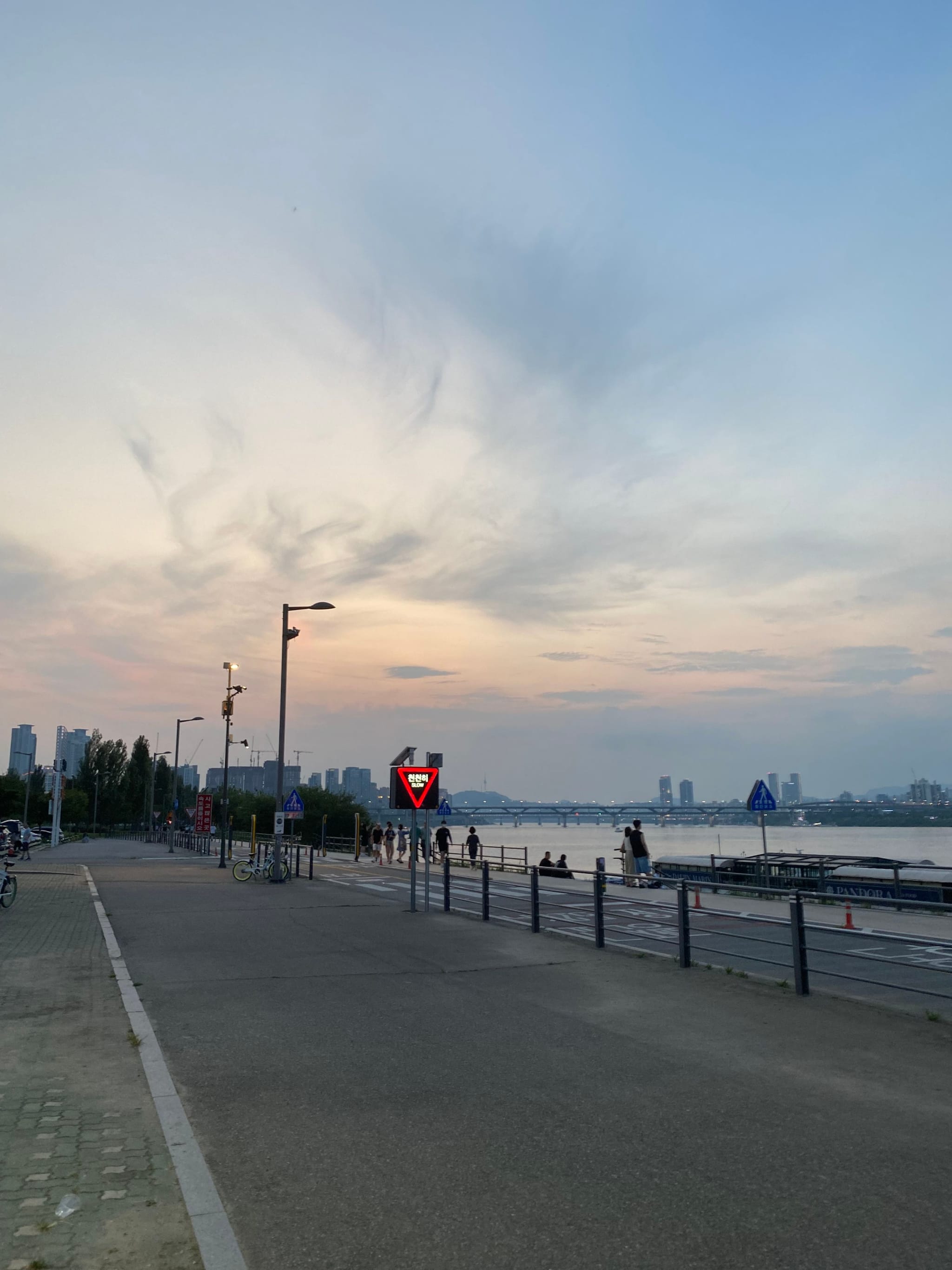 A riverside walkway at dusk with a few people strolling, streetlights, and a distant city skyline under a cloudy sky