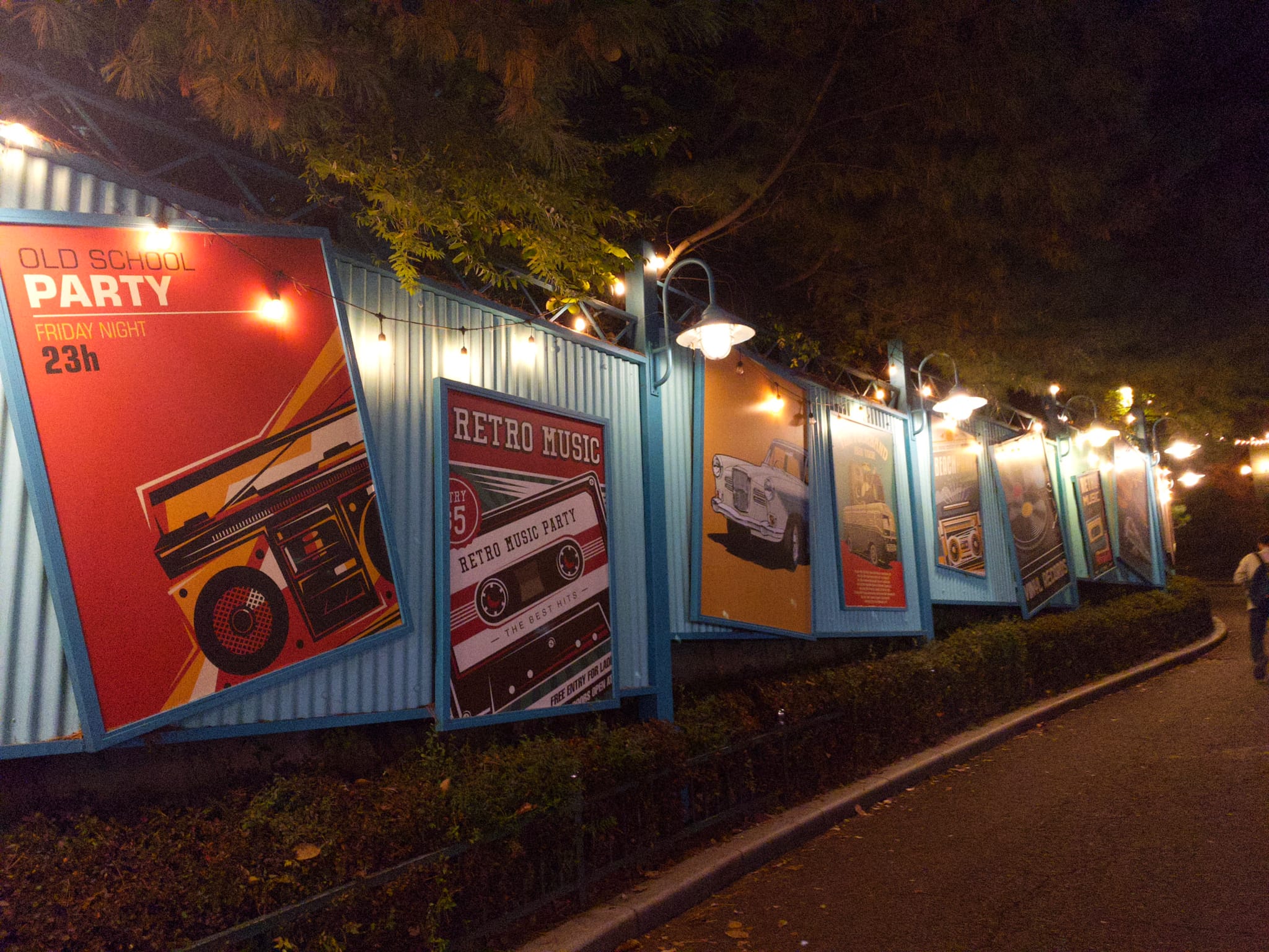 A row of colorful posters with retro designs is displayed on a blue wall, illuminated by overhead lights, along a pathway lined with trees