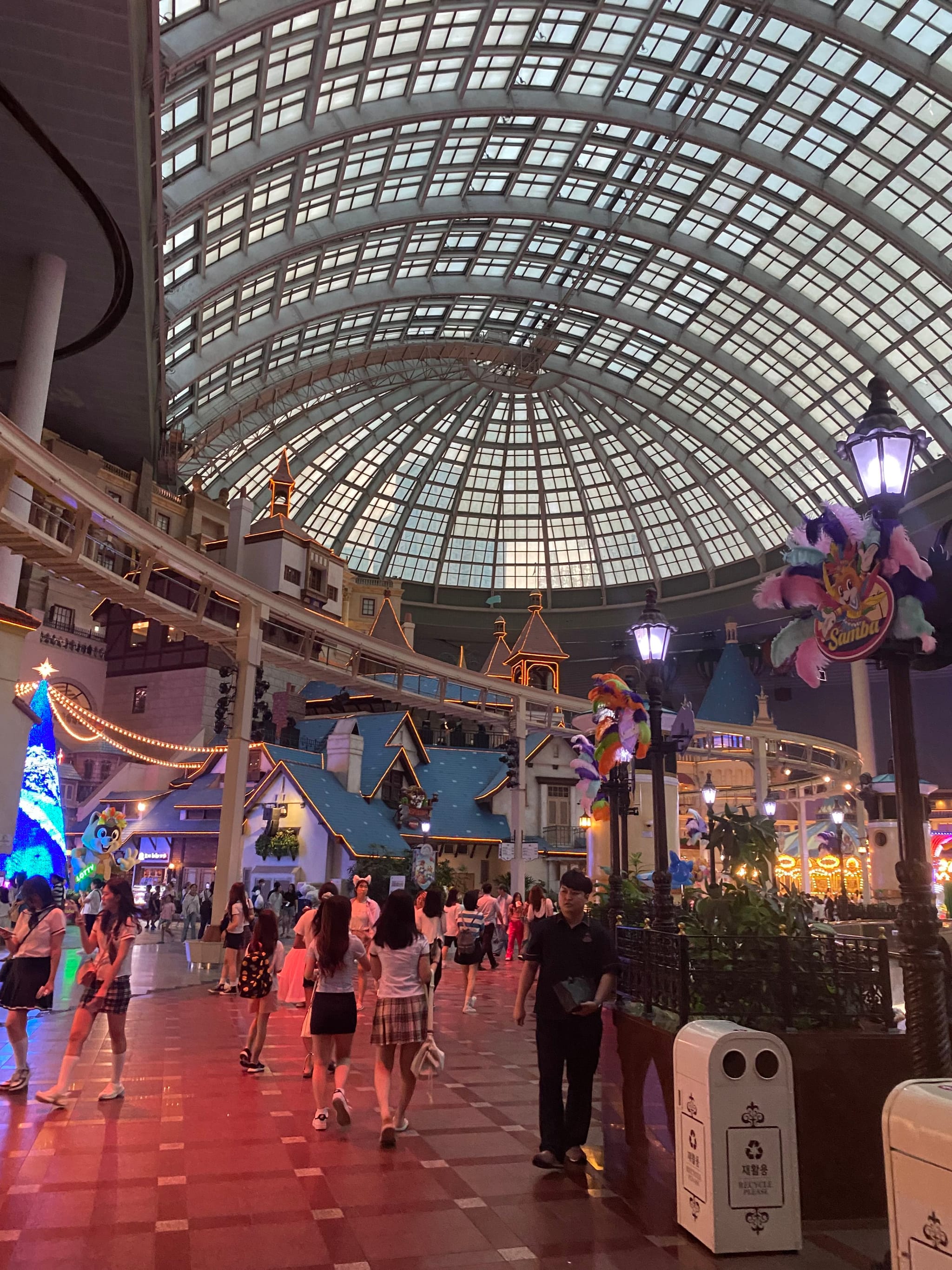 An indoor amusement park with a large glass dome ceiling, colorful decorations, and people walking around