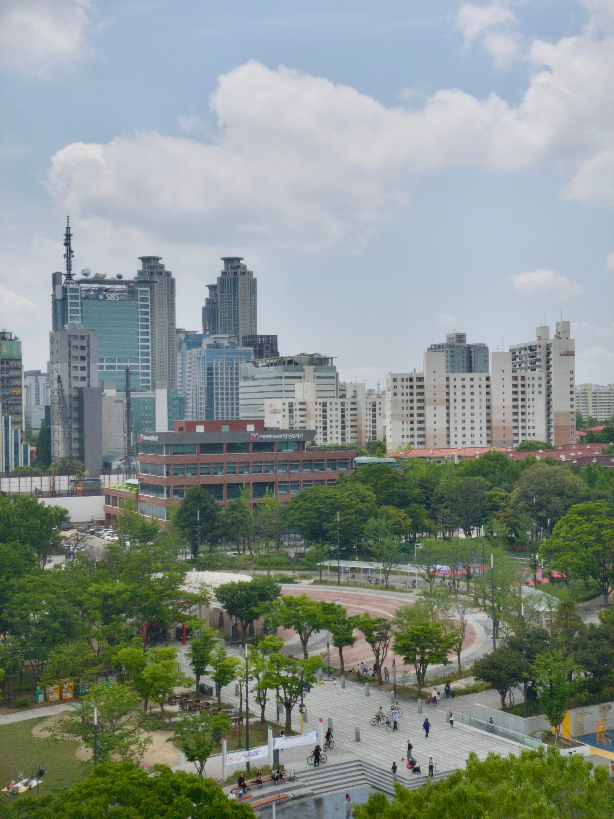 A cityscape featuring modern high-rise buildings in the background, a park with trees and pathways in the foreground, and a partly cloudy sky above