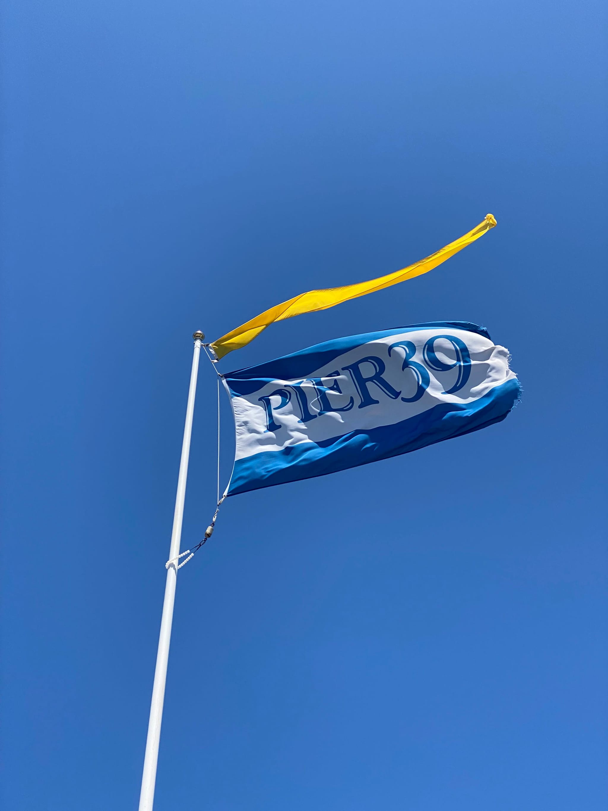 A blue flag with the word PIER 39 flutters on a pole against a clear blue sky, accompanied by a yellow pennant