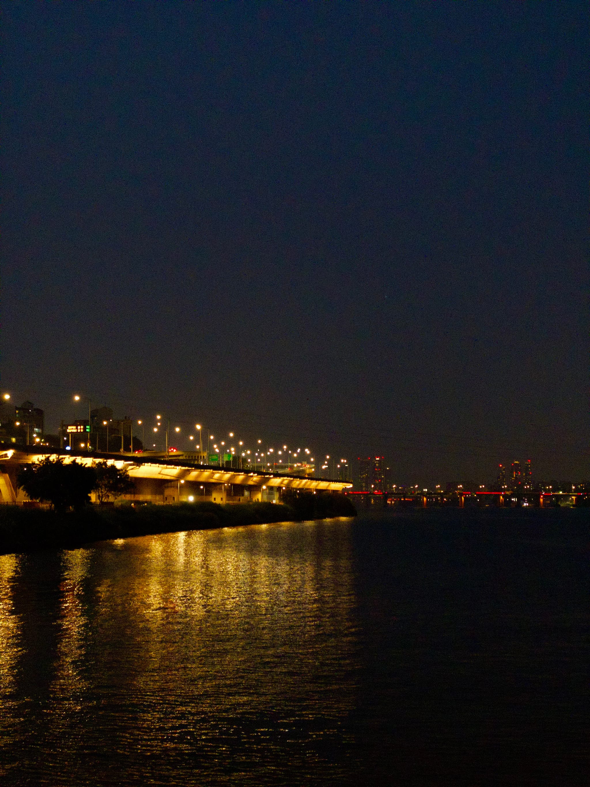 A bridge illuminated with lights spans across a body of water at night, with reflections shimmering on the surface