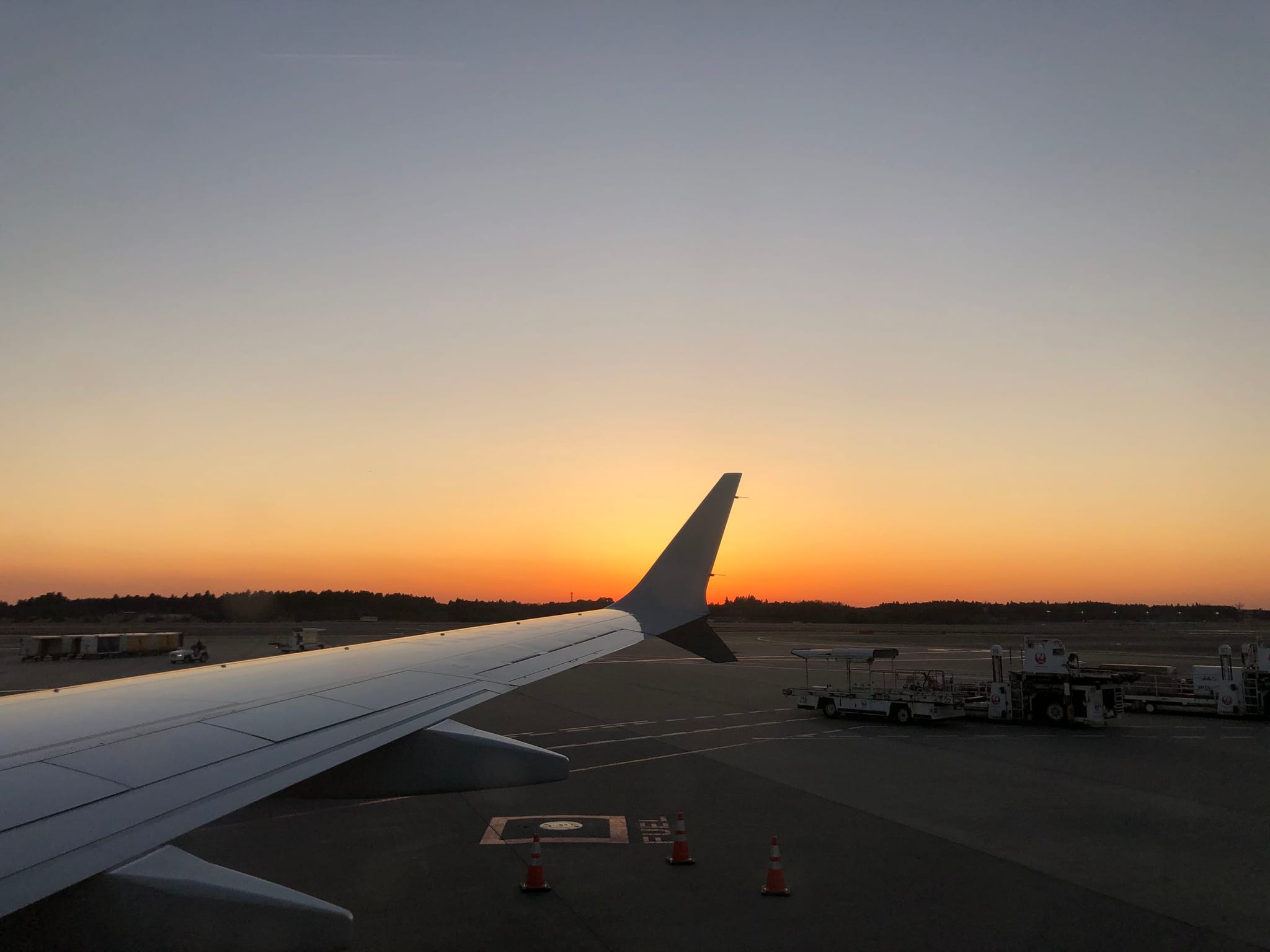 A view from an airplane window at sunset, with the wing visible and a clear sky transitioning from orange to blue. The airport tarmac and ground equipment are seen below
