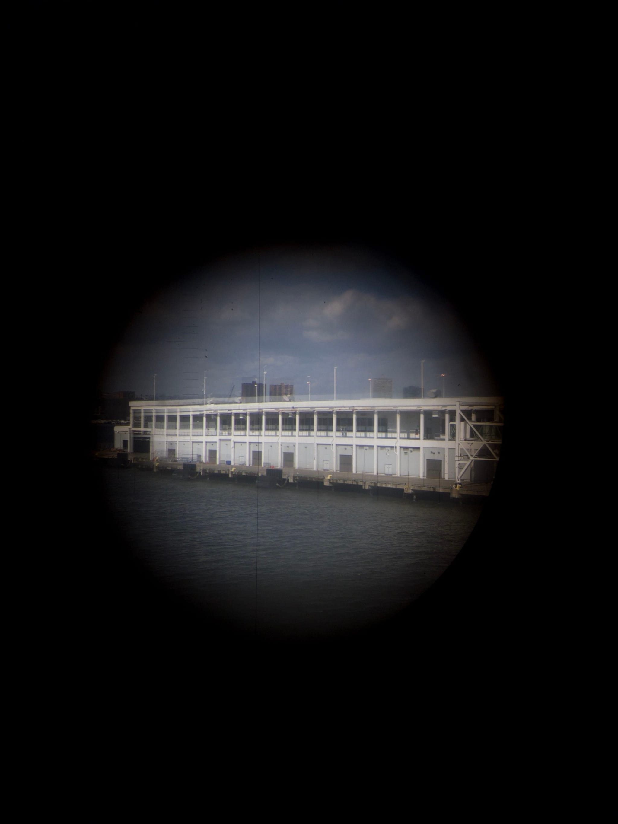 A white building with multiple windows is viewed through a circular lens, surrounded by darkness, with water in the foreground