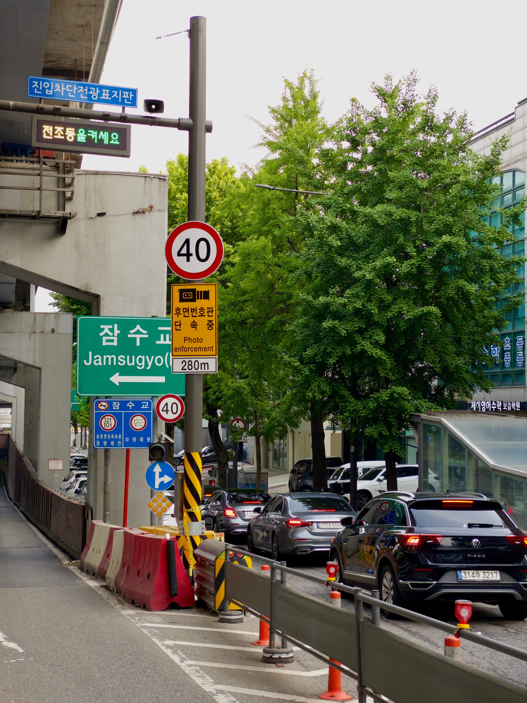 A street scene with traffic signs, including a speed limit of 40, and a green sign for Jamsugyo Bridge Cars are lined up on the road, and there are trees and buildings in the background