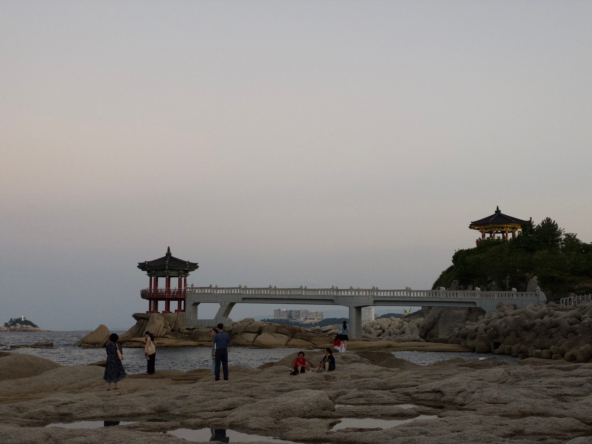 A rocky shore with people standing, featuring a traditional-style pavilion on the right and a small bridge leading to another structure on the left. Trees and rocks surround the area under a clear sky